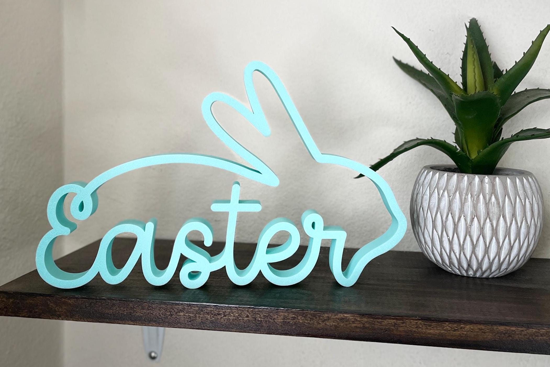 Light blue or teal 'Easter' sign shaped like a bunny, and a cross for the t. It sits on a shelf with a plant in the background.
